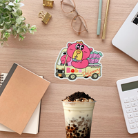 Sticker of a cartoon character in a van labeled 'Bobo Delivery' on a desk with a drink, notebook, and glasses.