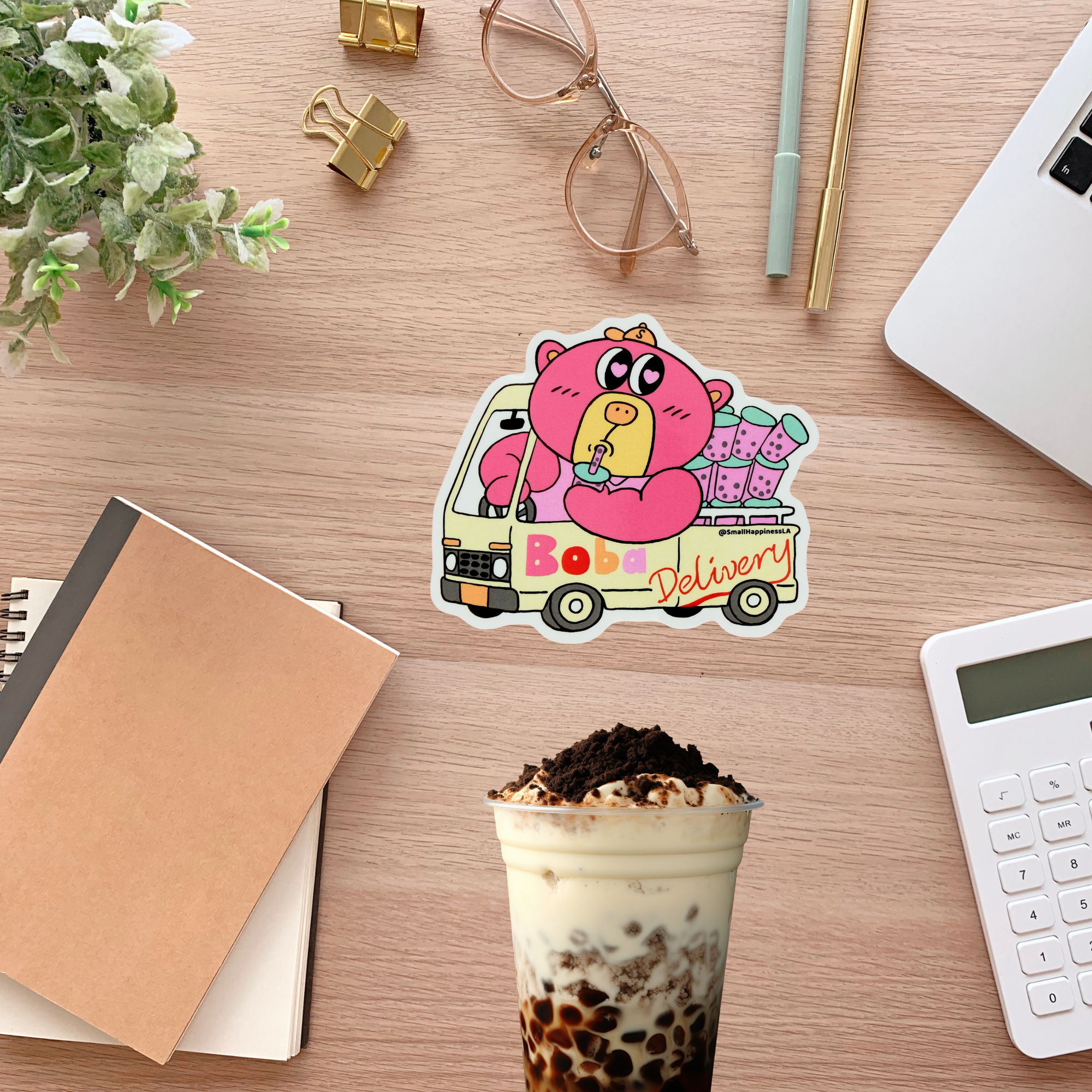Sticker of a cartoon character in a van labeled 'Bobo Delivery' on a desk with a drink, notebook, and glasses.