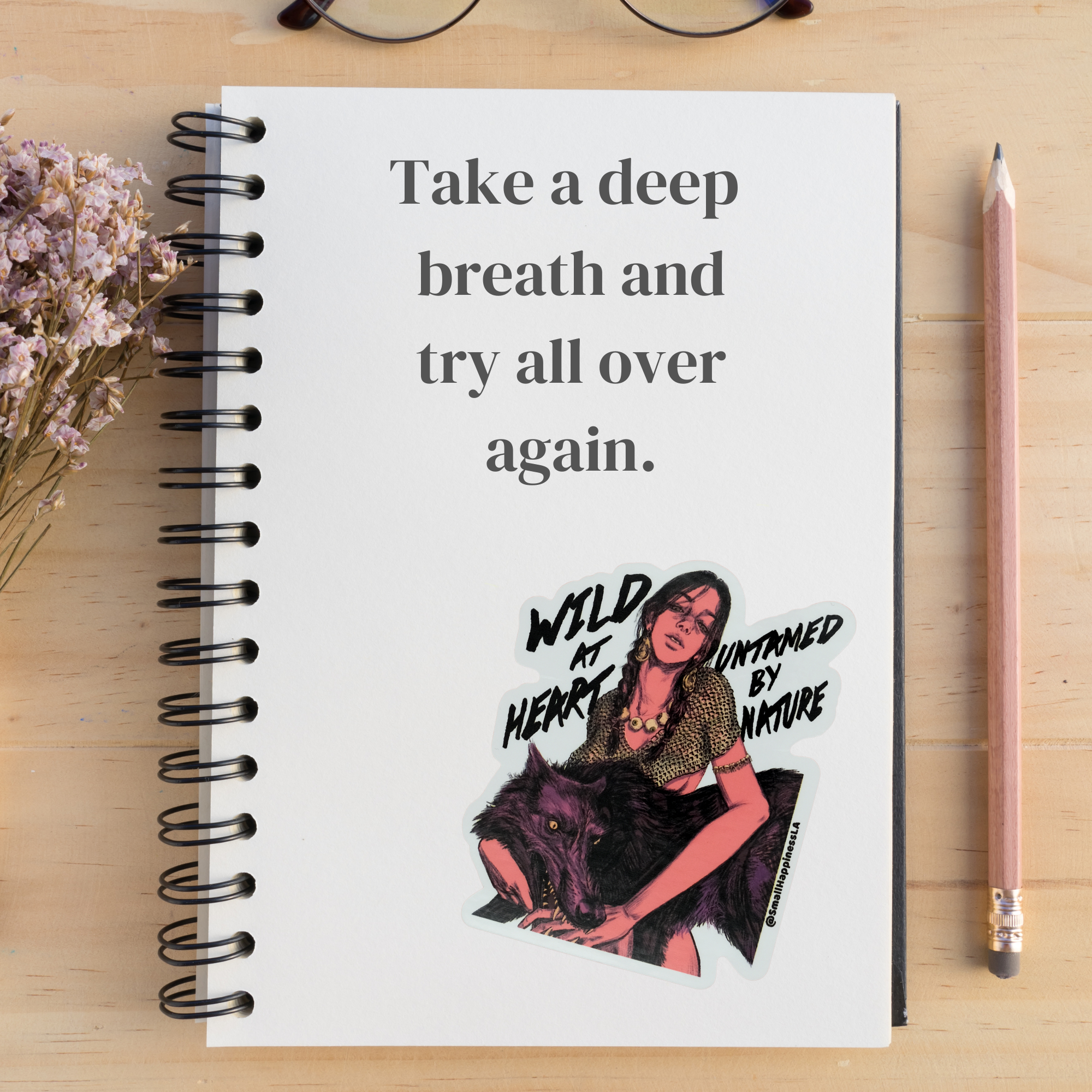 Notebook with motivational quote and illustration on a wooden surface with flowers and a pencil.