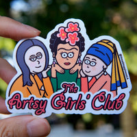 Artsy Girls Club vinyl sticker featuring cartoon versions of iconic women artists, including Mona Lisa, Frida Kahlo, and Girl with a Pearl Earring, on a pink background held by hand in natural day light