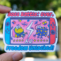 Boss Battle Frog vinyl sticker featuring two cartoon frogs battling on a handheld gaming console screen with the words “Boss Battle: Over” and “Freedom Unlocked” in bold, colorful text, held by hand in natural day light