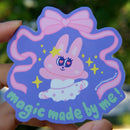 Hand holding a colorful sticker with a cartoon bunny character and text against a blurred background in natural lighting