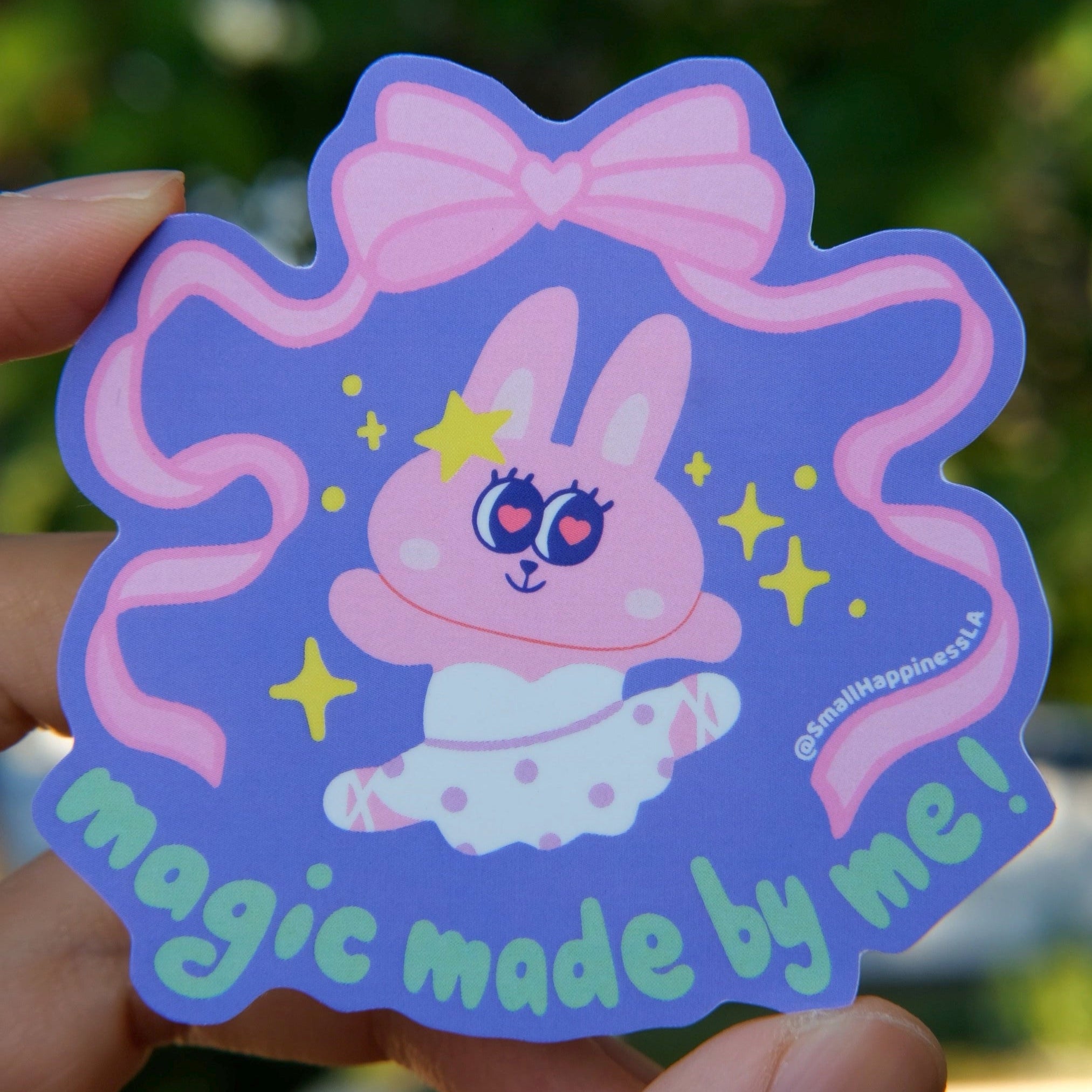 Hand holding a colorful sticker with a cartoon bunny character and text against a blurred background in natural lighting