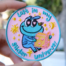 Colorful in my sticker universe-Mochi the frog embroidery patch held by a hand with a blurred natural background.
