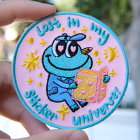Colorful in my sticker universe-Mochi the frog embroidery patch held by a hand with a blurred natural background.
