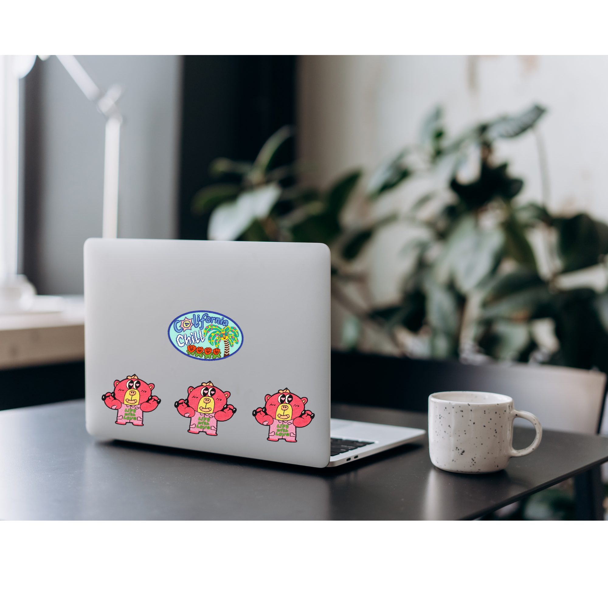 Laptop with stickers on a desk next to a mug, with a blurred plant in the background
