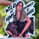 Vinyl sticker held by hand in natural day light featuring a fierce young woman with braids and jewelry holding a black wolf, with bold text that reads “Wild at Heart, Untamed by Nature,” designed in a dark, moody art style by SmallHappinessLA