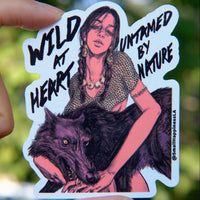 Vinyl sticker held by hand in natural day light featuring a fierce young woman with braids and jewelry holding a black wolf, with bold text that reads “Wild at Heart, Untamed by Nature,” designed in a dark, moody art style by SmallHappinessLA