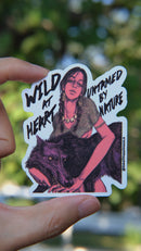 Vinyl sticker held by hand in natural day light featuring a fierce young woman with braids and jewelry holding a black wolf, with bold text that reads “Wild at Heart, Untamed by Nature,” designed in a dark, moody art style by SmallHappinessLA