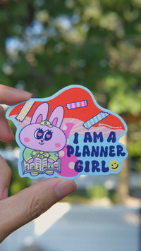 Planner Girl vinyl sticker featuring a cute pink bunny reading a planner and the phrase "I am a planner girl" in bold letters, held by hand in natural day light, designed by SmallHappinessLA
