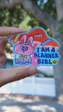 Video of Planner girl embroidery patch held in hand, showing size, texture, stitching details, and fabric under sunlight.
