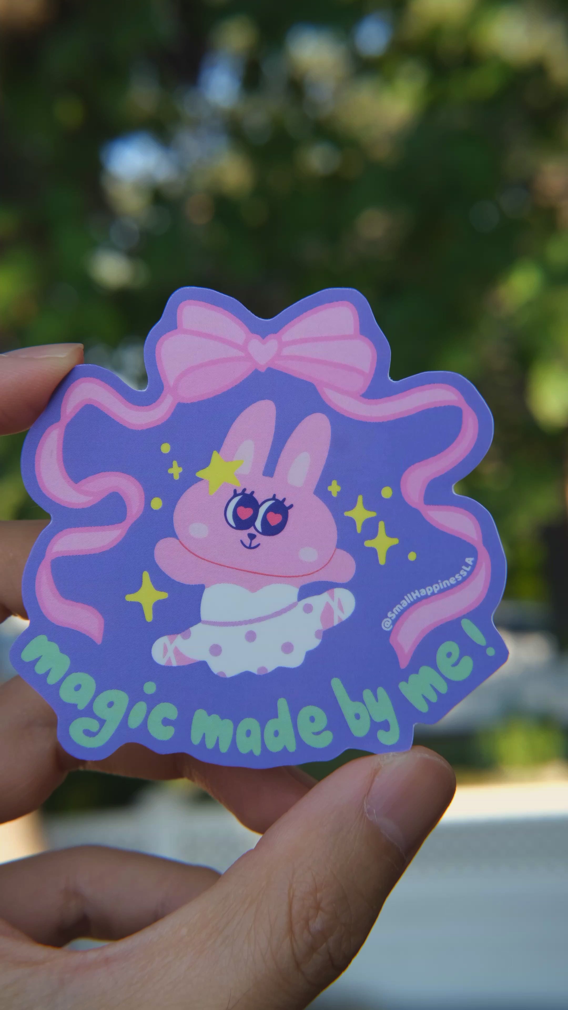Hand holding a colorful sticker with a cartoon bunny character and text against a blurred background in natural lighting
