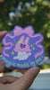 Hand holding a colorful sticker with a cartoon bunny character and text against a blurred background in natural lighting