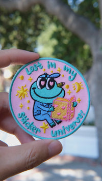 Video of in my sticker universe-Mochi the frog embroidery patch held in hand, showing size, texture, stitching details, and fabric under sunlight.