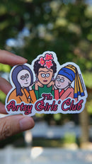 Artsy Girls Club vinyl sticker featuring cartoon versions of iconic women artists, including Mona Lisa, Frida Kahlo, and Girl with a Pearl Earring, on a pink background held by hand in natural day light
